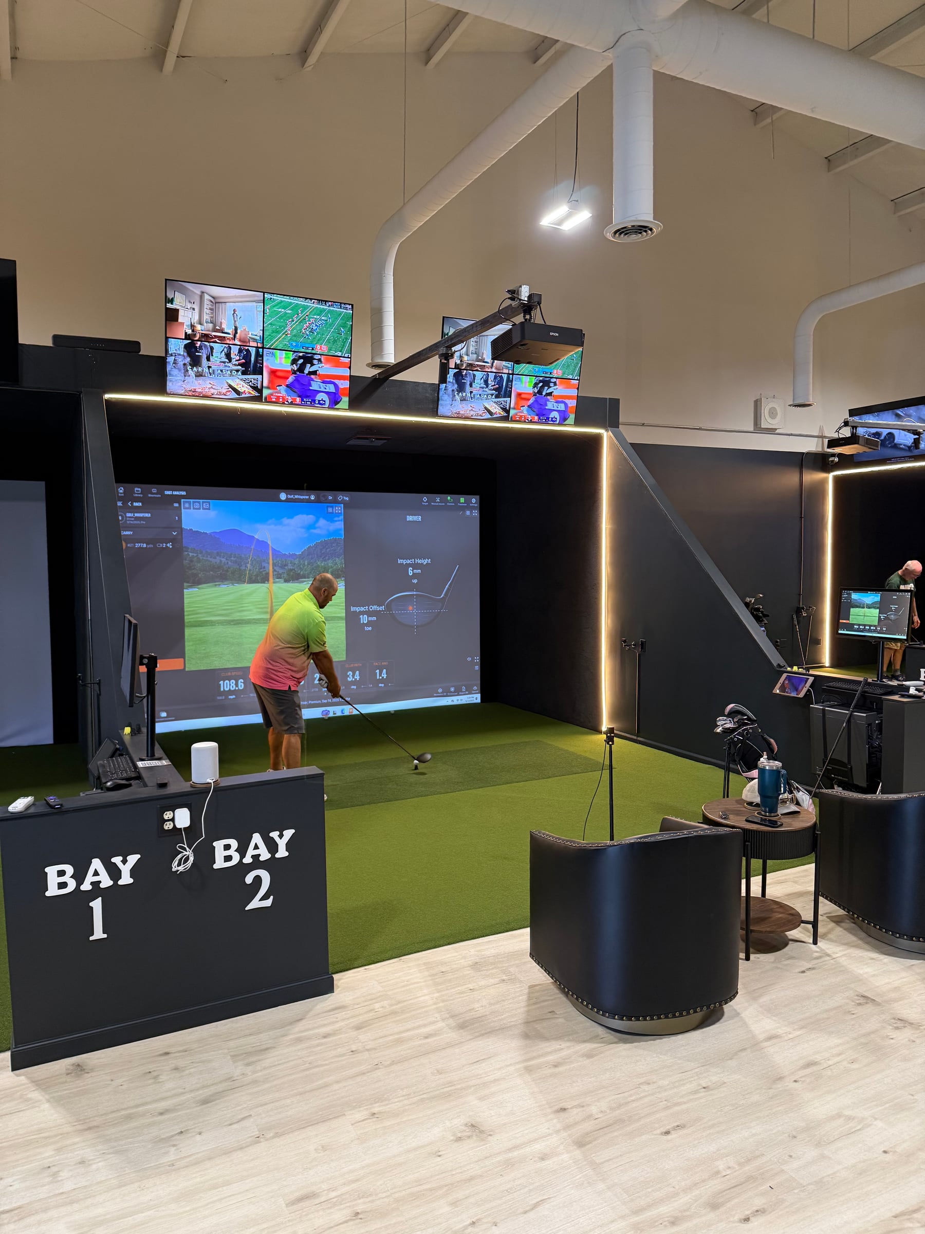 Ohio Golf Lounge — BAY 1 / BAY 2 signage on a black podium, LED-strip-lit black bays, pale timber floor, golf